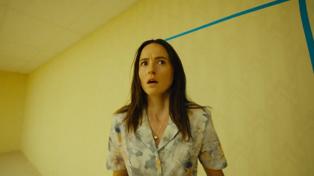 A frightened woman alone in one of the empty yellow rooms of the A24 horror film Backrooms